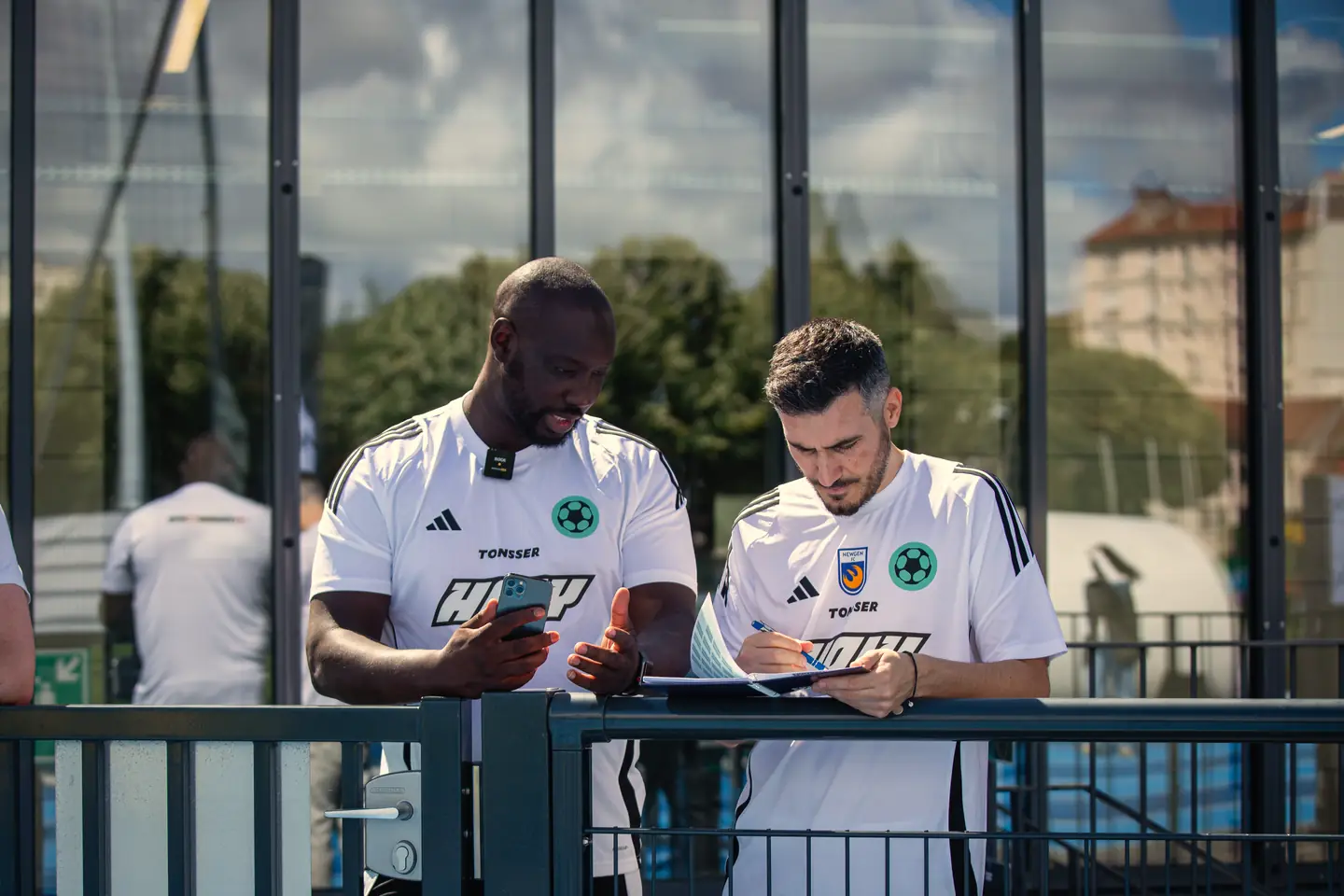 Two Tonsser coaches by the pitch, one checking a phone, the other taking notes.